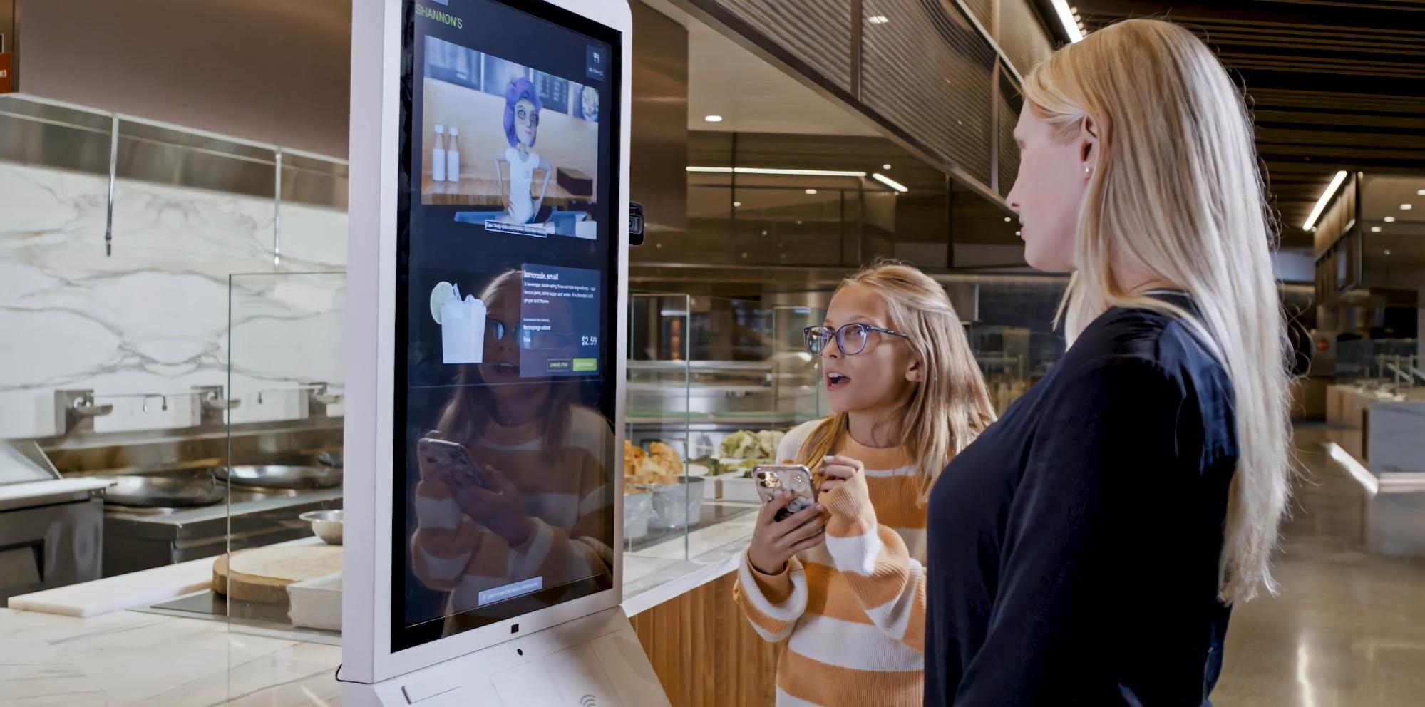 Photo shows a woman and child interact with an avatar in a restaurant ordering kiosk.