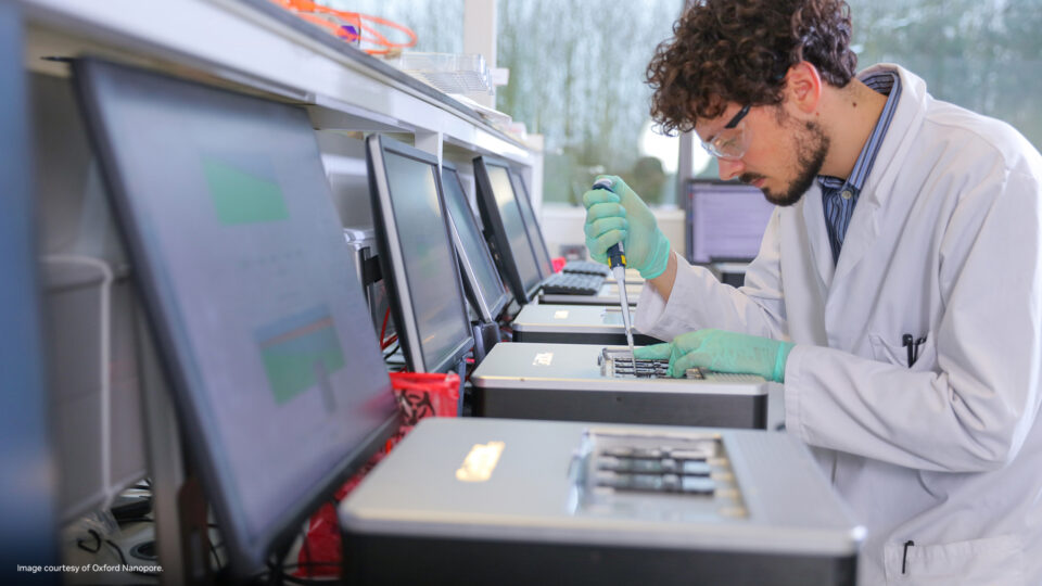 Photo of a scientist in a lab coat and holding a pipette working over a row of machines.