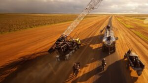Post-visualization still from Mad Max: Furiosa. A close-up view of a desert chase scene after a disaster. The scene has modified vehicles, including a big tanker truck, a crane-like vehicle, motorbikes, and a pickup truck driving fast across a dusty, reddish-brown road under a dramatic, cloudy sky.