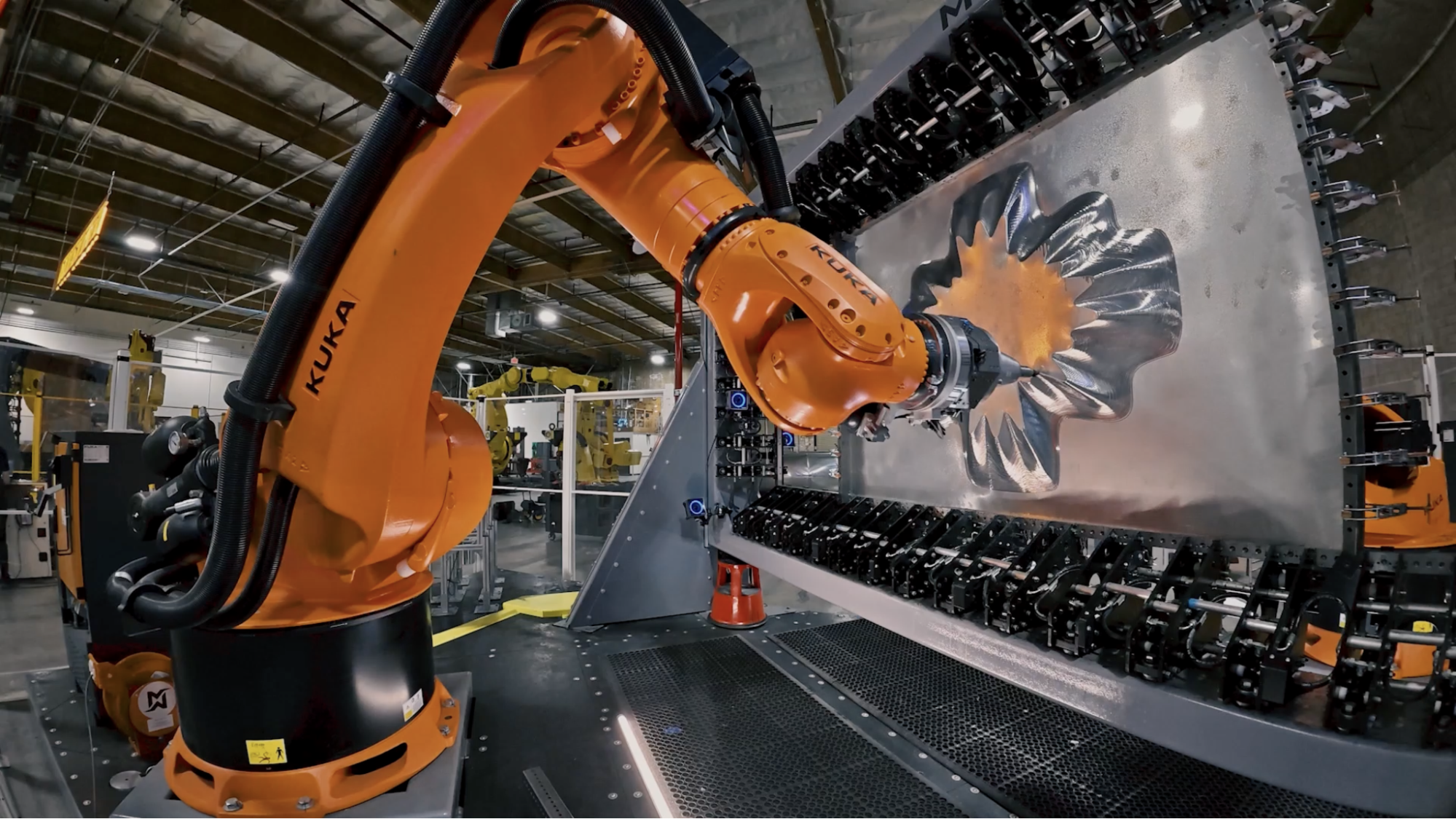 An image of a piece of metallic sculpture art, with a large orange robotic arm in the foreground in front of a large sheet of metal the robotic arm has been carved into a shape that looks like a flower or four-leaf clover.