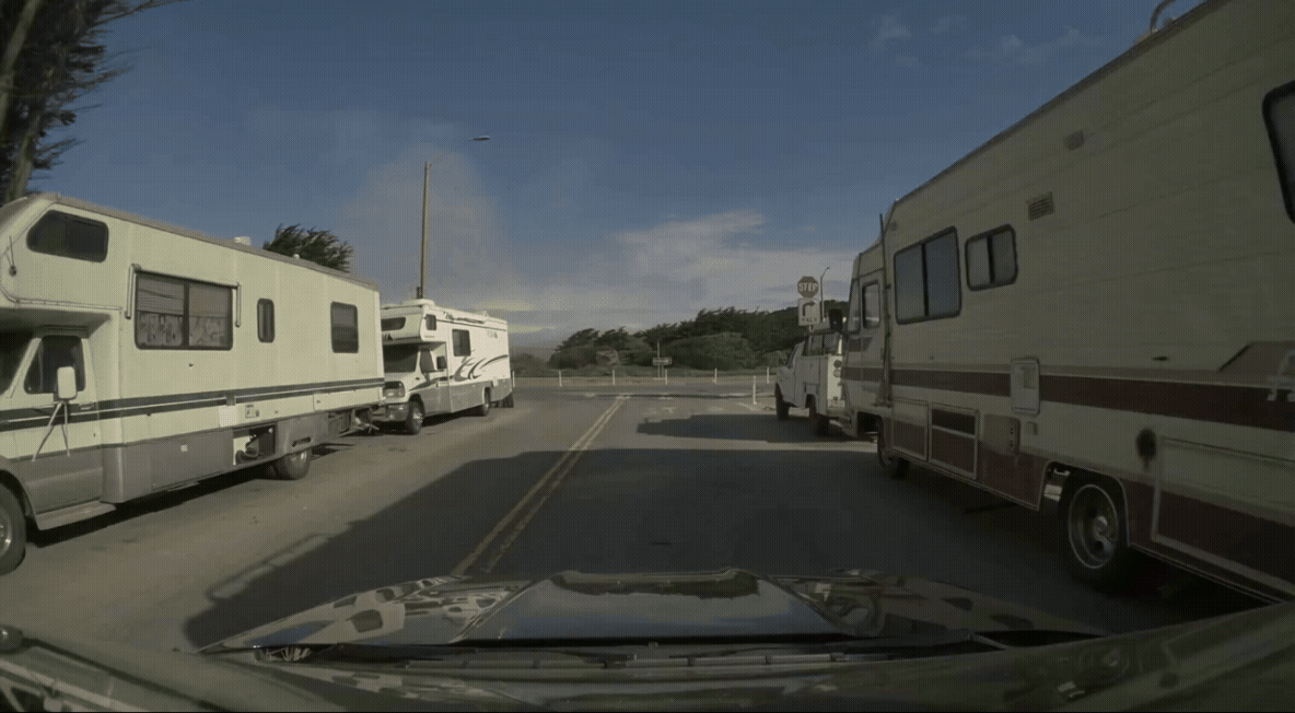 GIF of a car driving down a street with parked vehicles and a visible Stop sign near a curve.